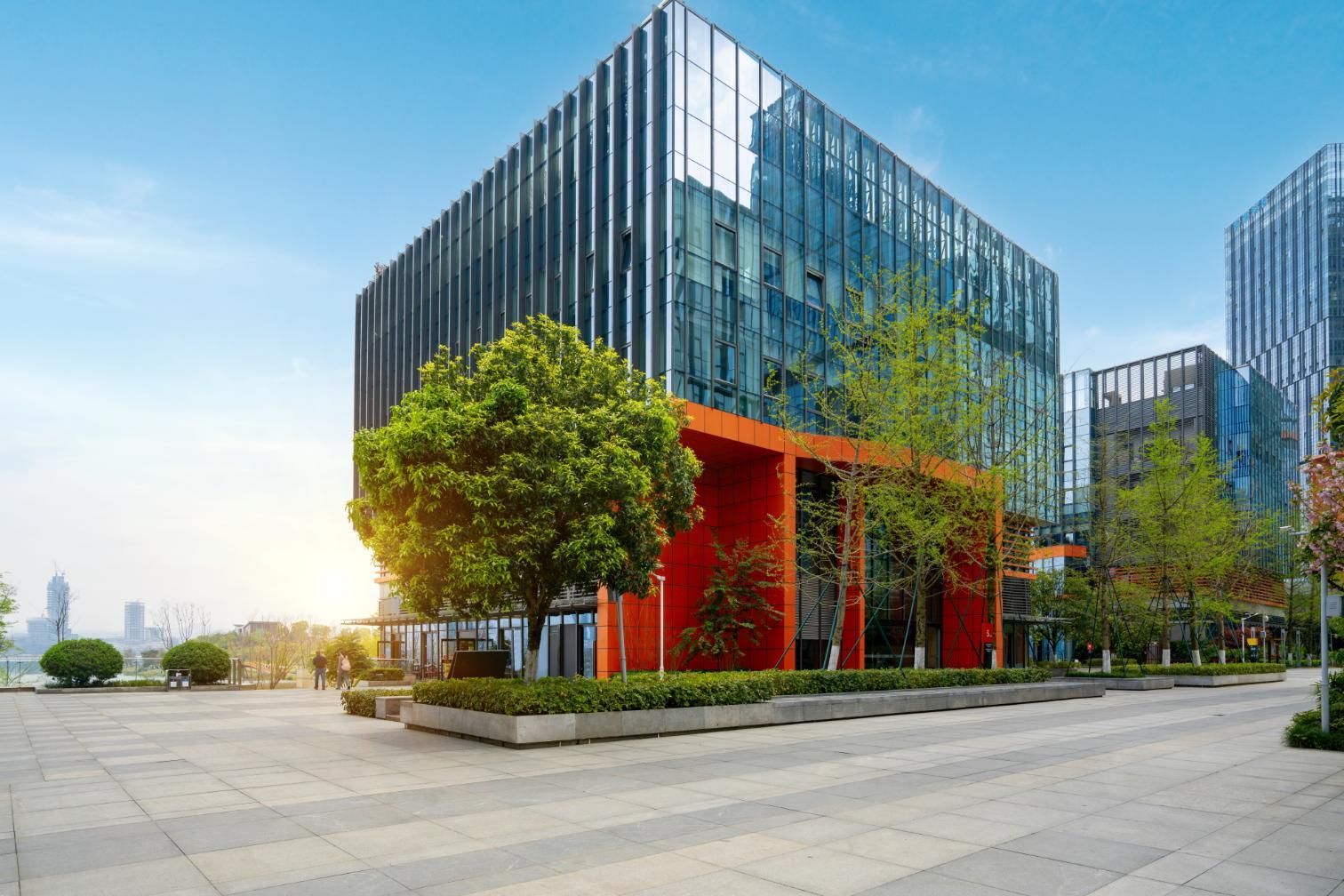 Modern glass office building with trees, orange accents, and a paved walkway.