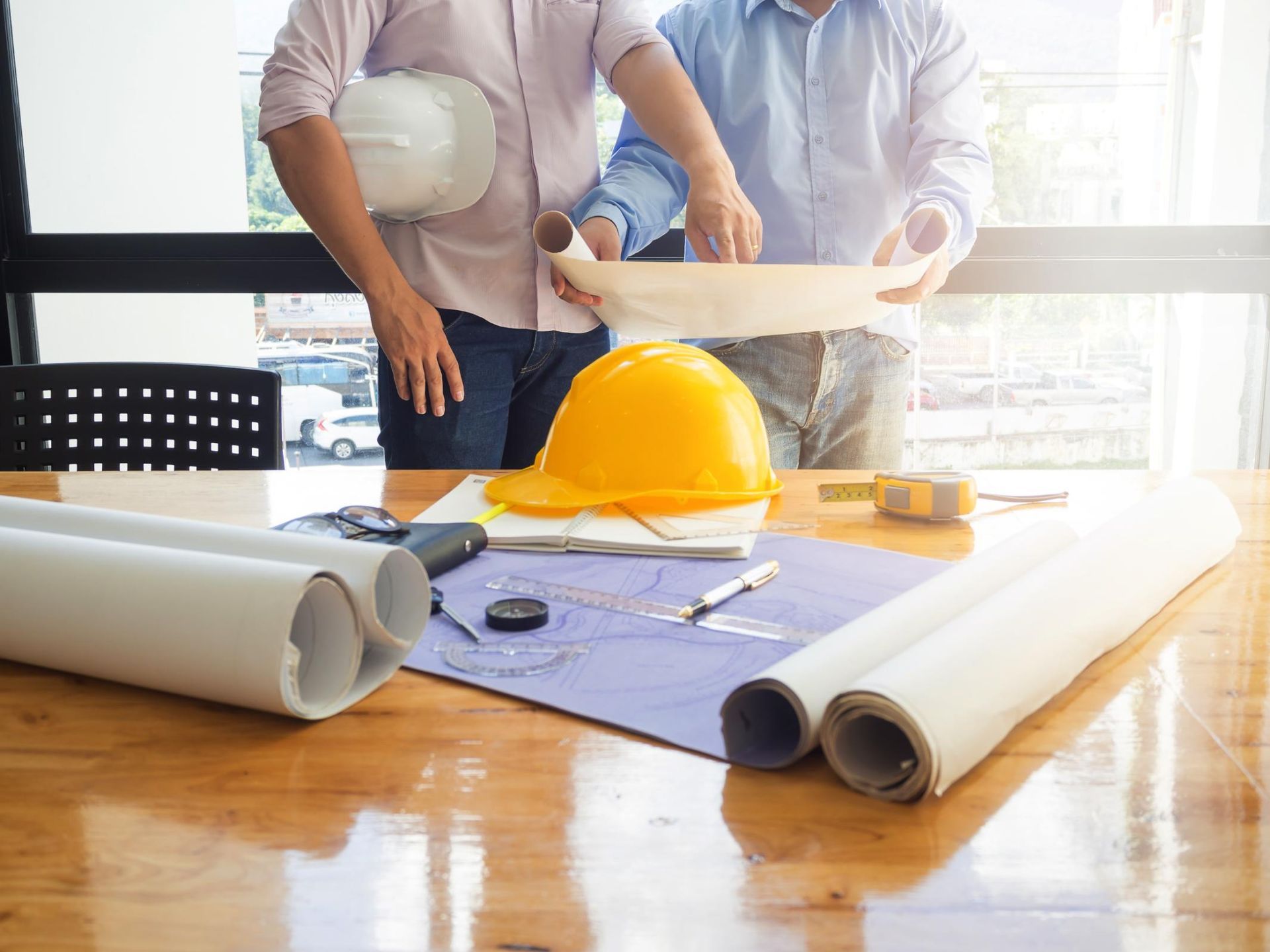 Two construction workers reviewing blueprints at a table with tools and safety helmets.