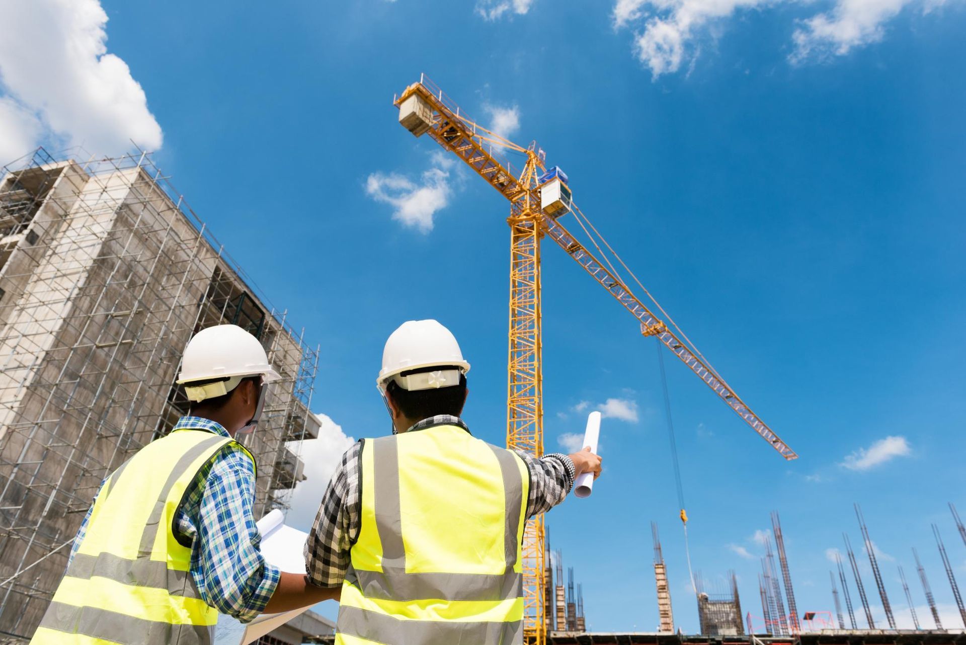 Two construction workers in hard hats and vests examine plans at a construction site with a crane.