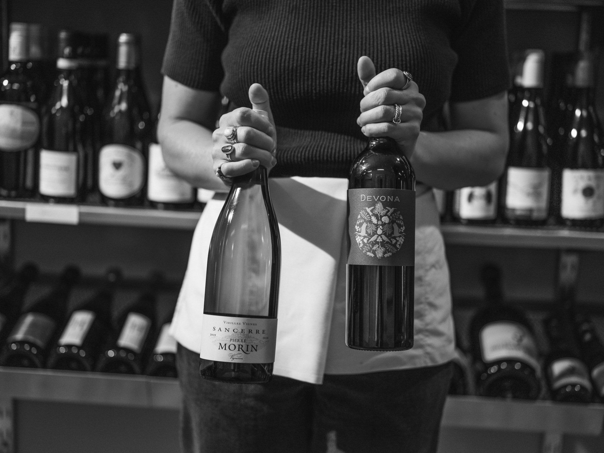 A woman is holding a bottle of wine in front of a shelf of wine bottles.