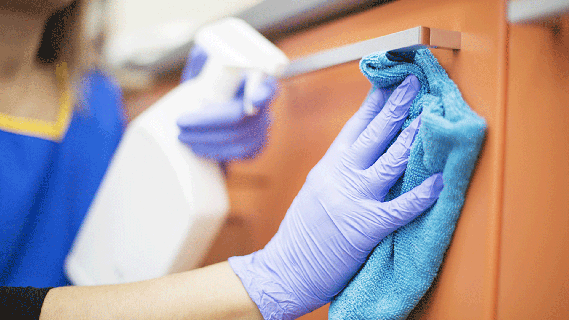 Person wearing gloves spraying cleaner and wiping down a cabinet surface with a microfibre cloth