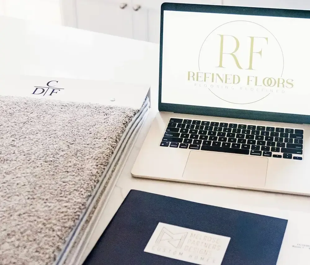 A laptop displaying the Refined Floors logo, a carpet sample, and a dark folder arranged on a white workspace.