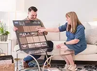 A person shows a flooring sample board to another person while they sit on a couch in a brightly lit living room.