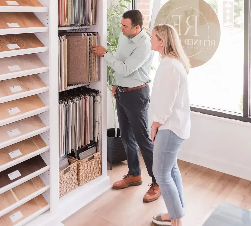 A man and woman examine fabric samples at a design showroom, looking at hanging textiles displayed on shelves.