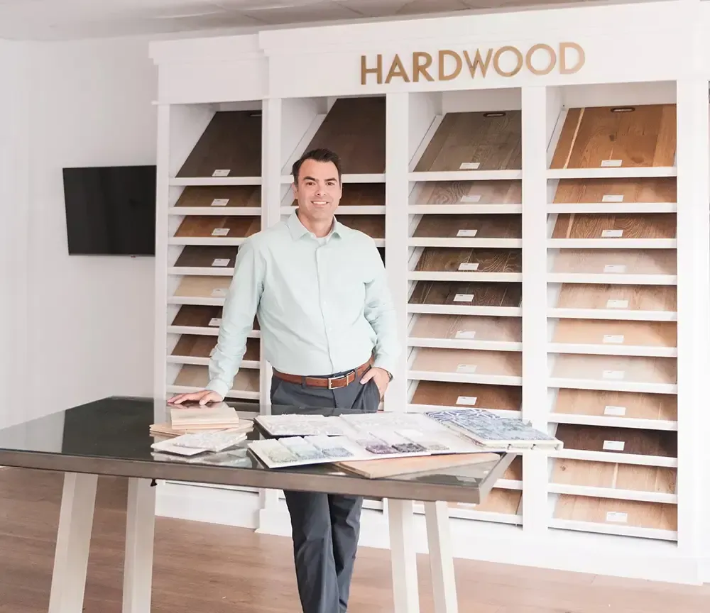 A person smiles while standing at a table in a hardwood flooring showroom, with shelves of wood samples in the background.