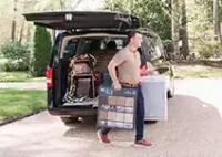 A person in a tan polo and jeans walks from a van, carrying a brown carpet sample board and a white item on a driveway.