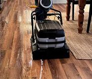 A person uses a walk-behind floor scrubbing machine to clean wooden flooring in a home.