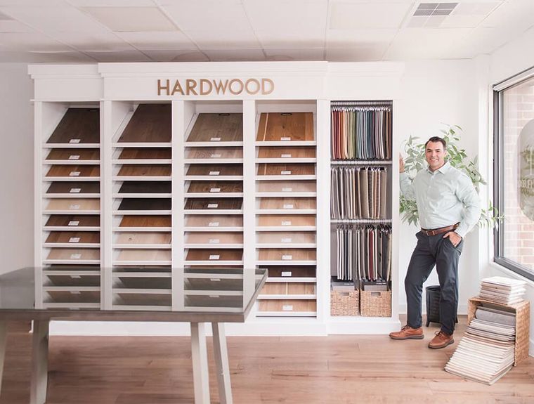 A professional stands in a showroom by walls of wood flooring samples and fabric swatches near a table and window.