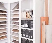 A showroom display features slanted shelves with wood flooring samples, hanging fabric swatches, and rows of black binders.