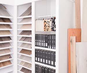 A showroom display features slanted shelves with wood flooring samples, hanging fabric swatches, and rows of black binders.