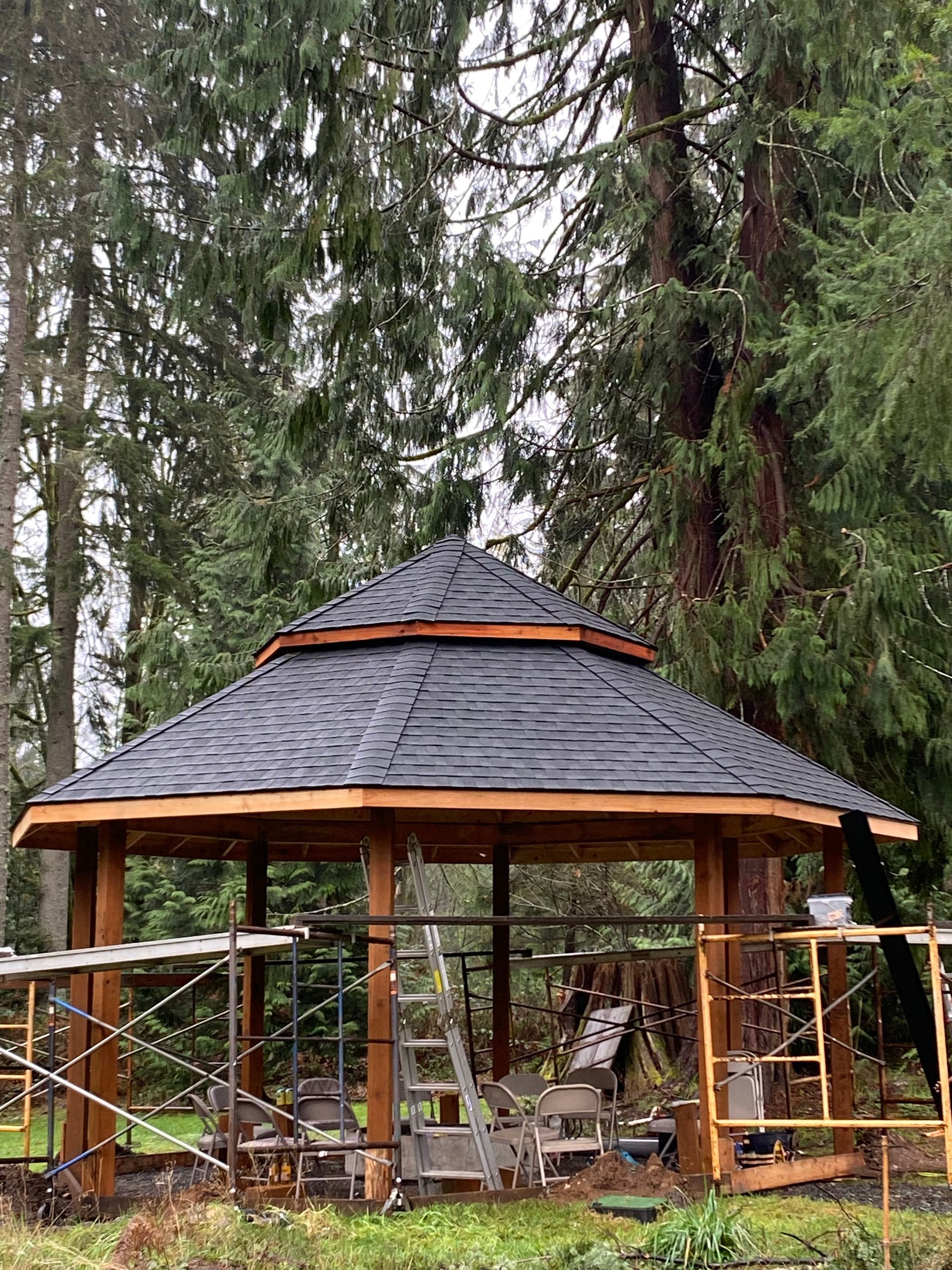 Partially built wooden gazebo with dark shingled roof, surrounded by trees and scaffolding.