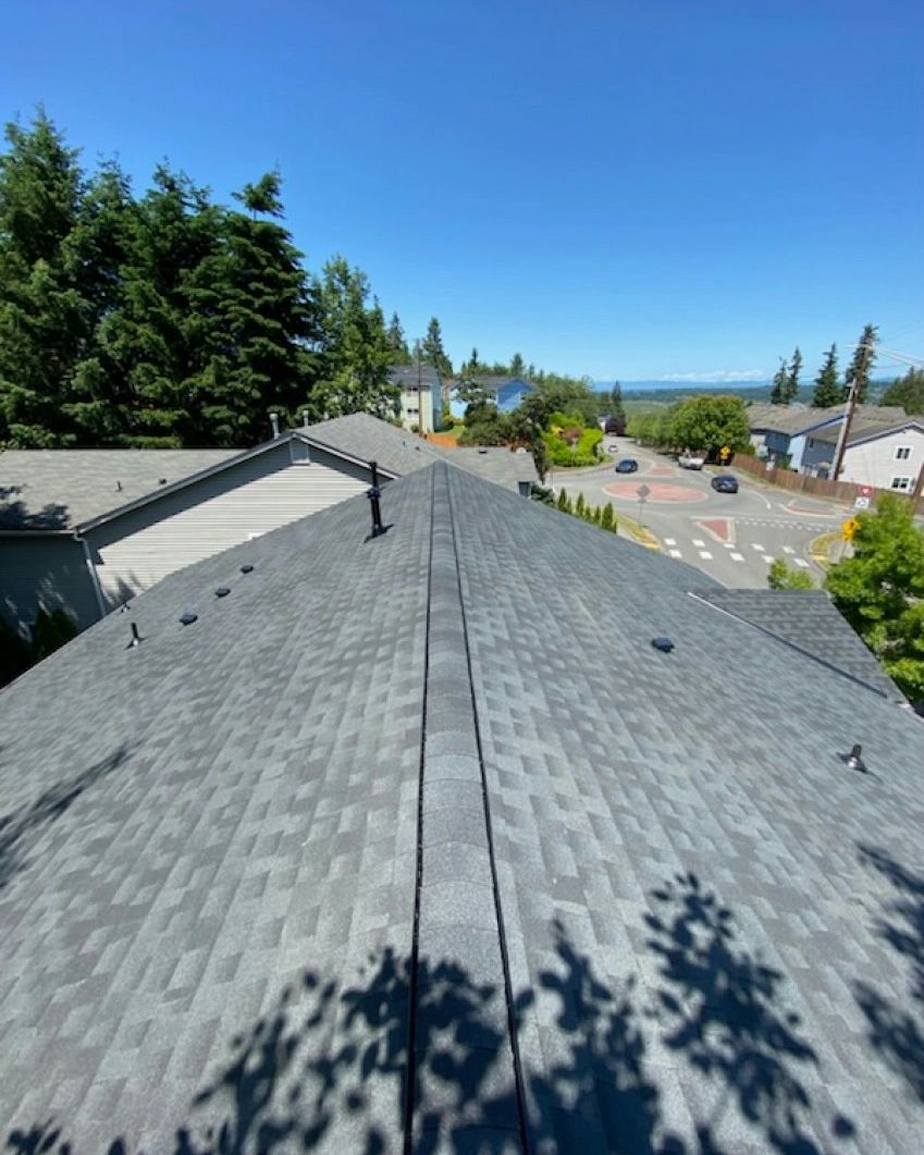 A rooftop with grey shingles, two vent pipes, and a view of trees, roads, and a blue sky.