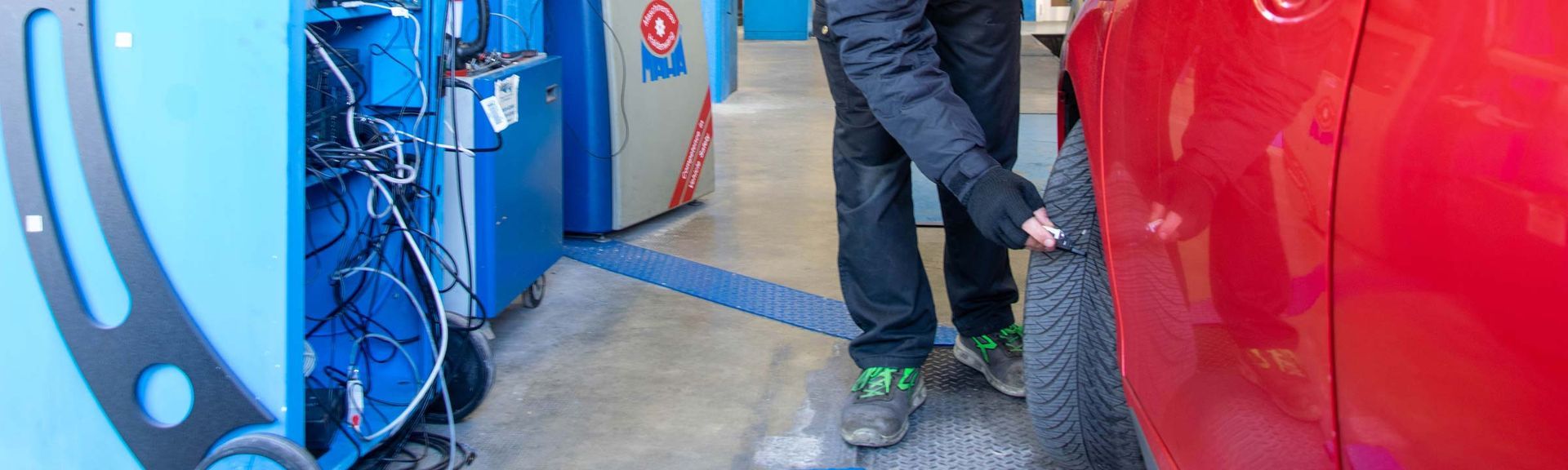 Una persona controlla gli pneumatici di un'auto rossa in un'autofficina. Sono visibili le attrezzature blu e il pavimento in cemento.