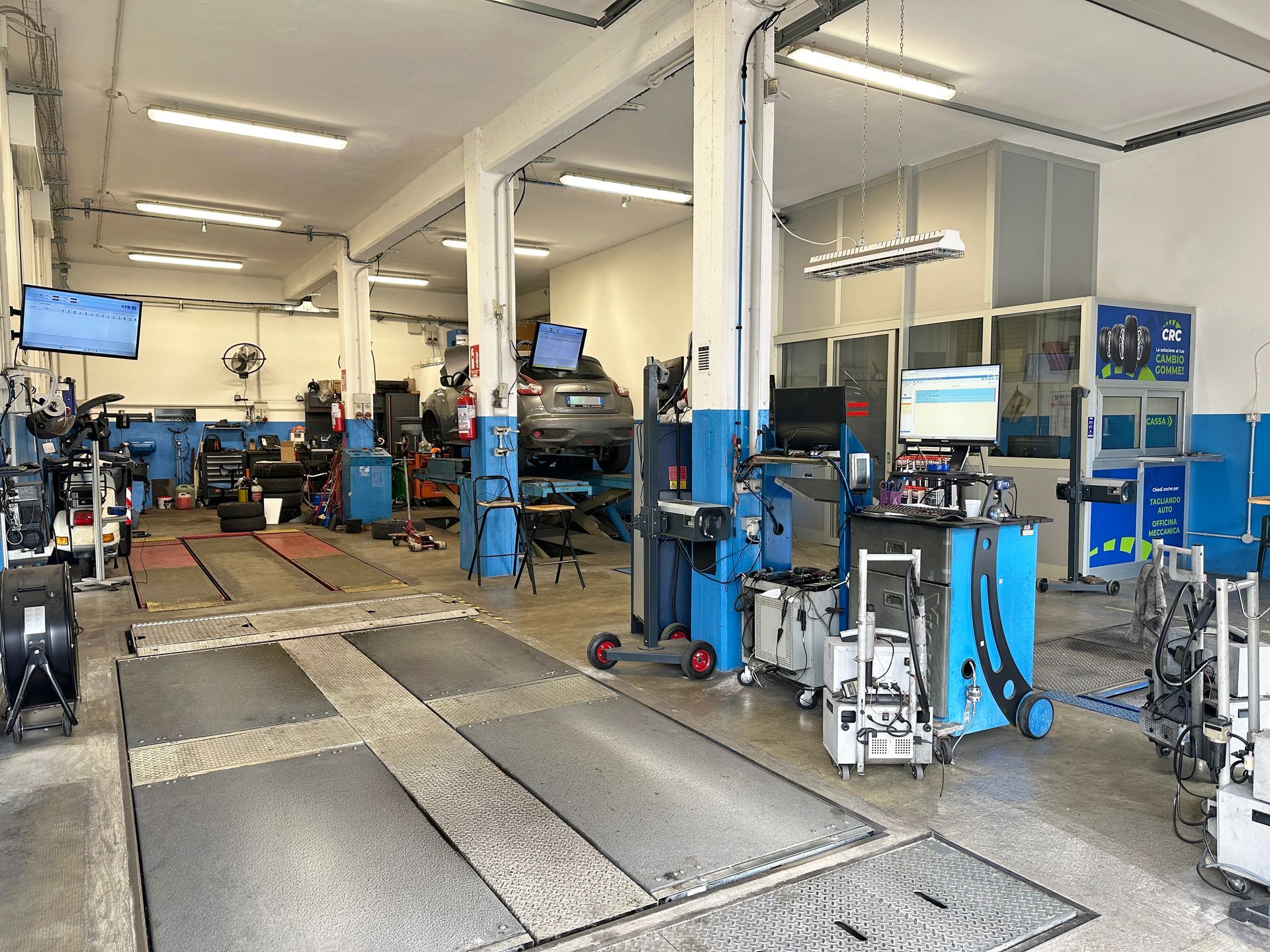 Un'auto su un ponte sollevatore in un'officina. Un meccanico con la mascherina lavora sotto. Sono presenti i colori blu e bianco.