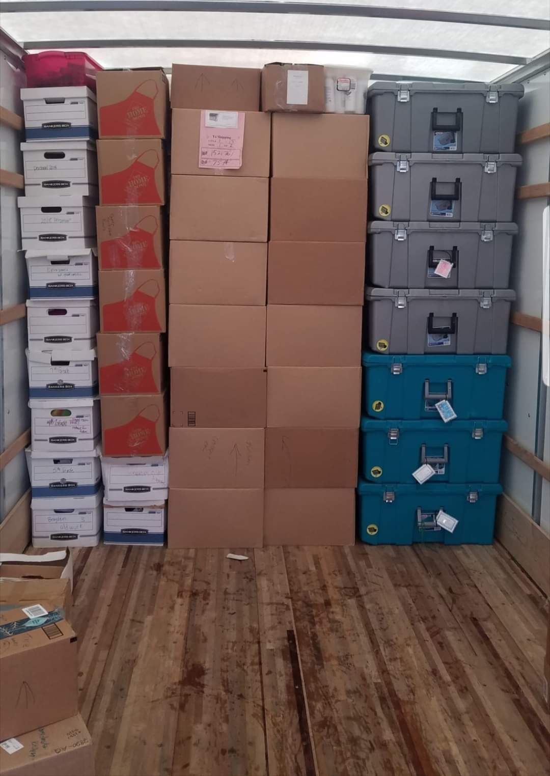 Boxes and bins stacked inside a truck: cardboard, gray, teal, and red. Wooden floor, white ceiling.