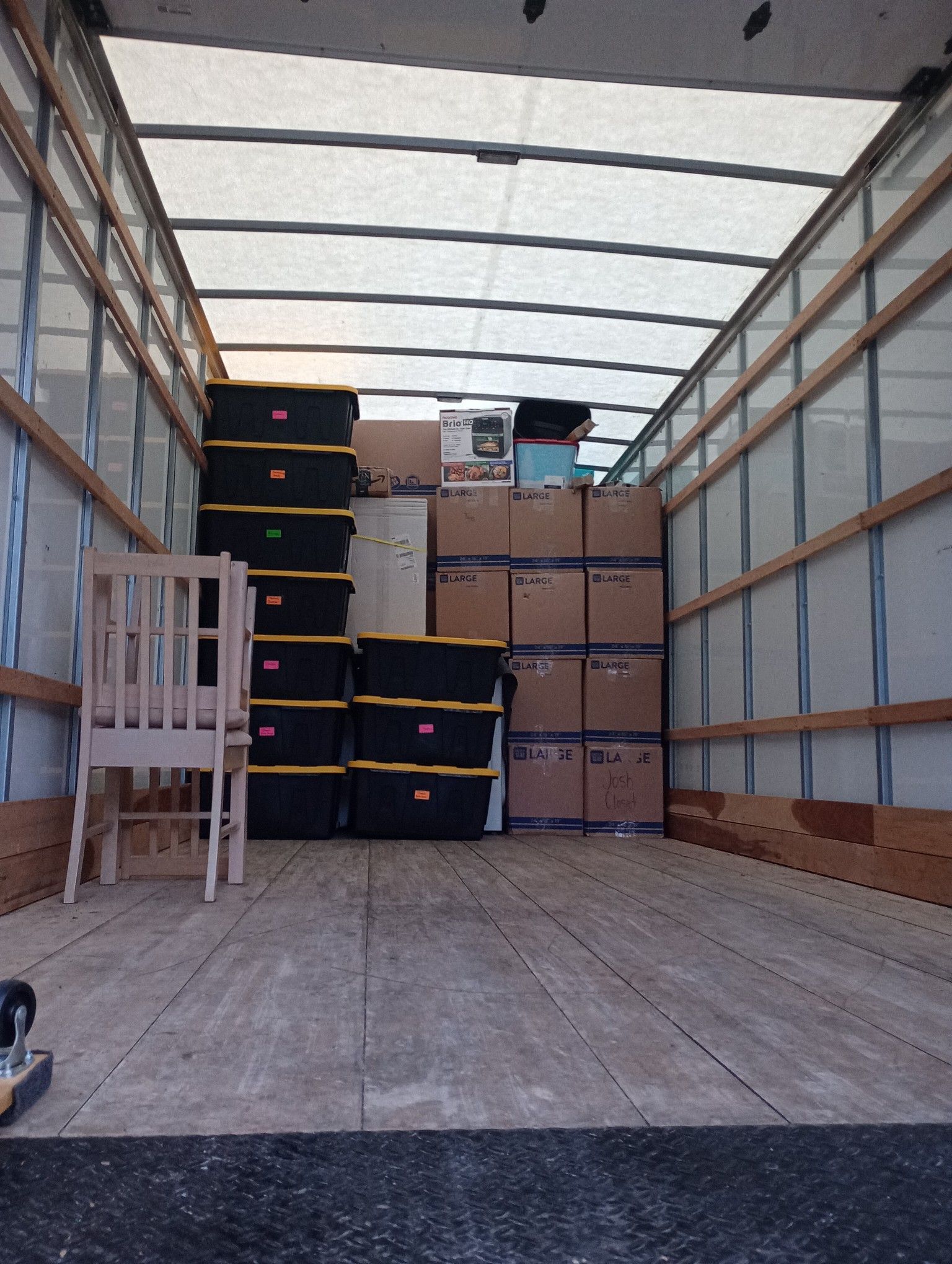 Inside a moving truck packed with black bins, boxes, and a wooden chair.