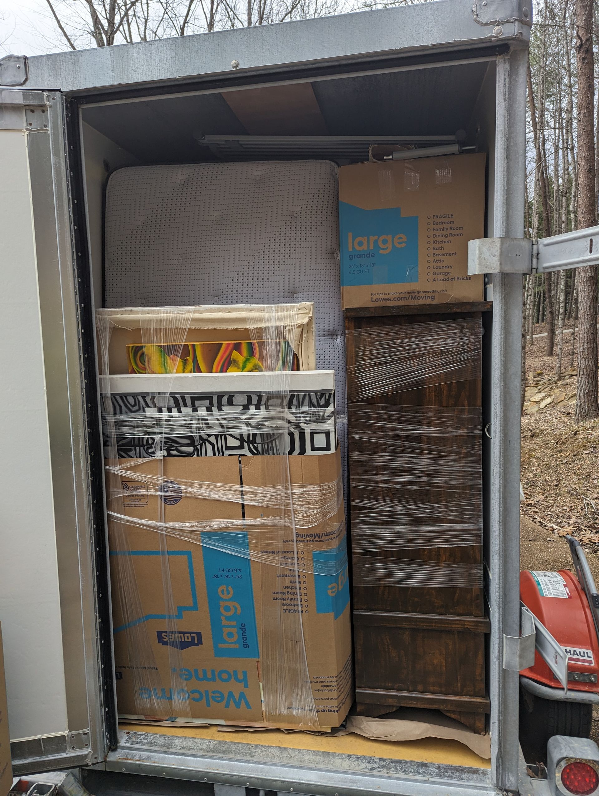 A trailer packed with moving boxes, a mattress, and furniture. The trailer's door is open.