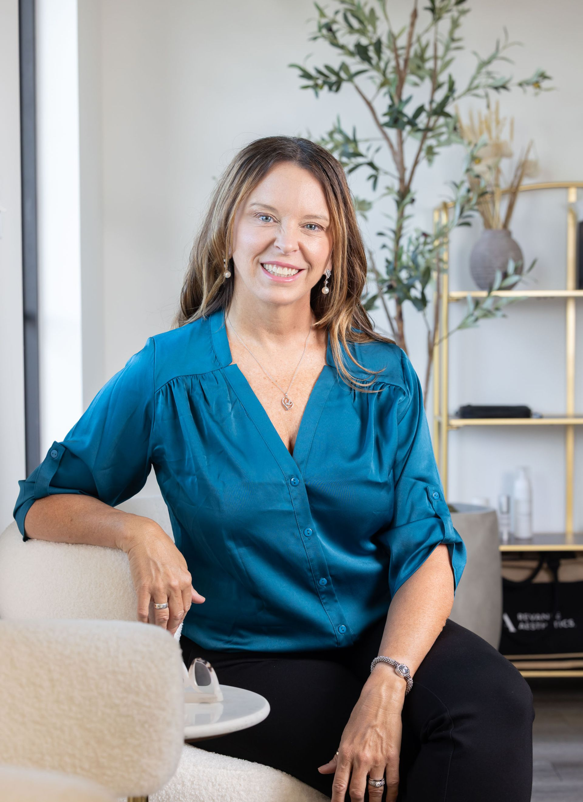 Woman in a teal blouse smiling while seated in a chair, with a plant and a shelf in the background office.