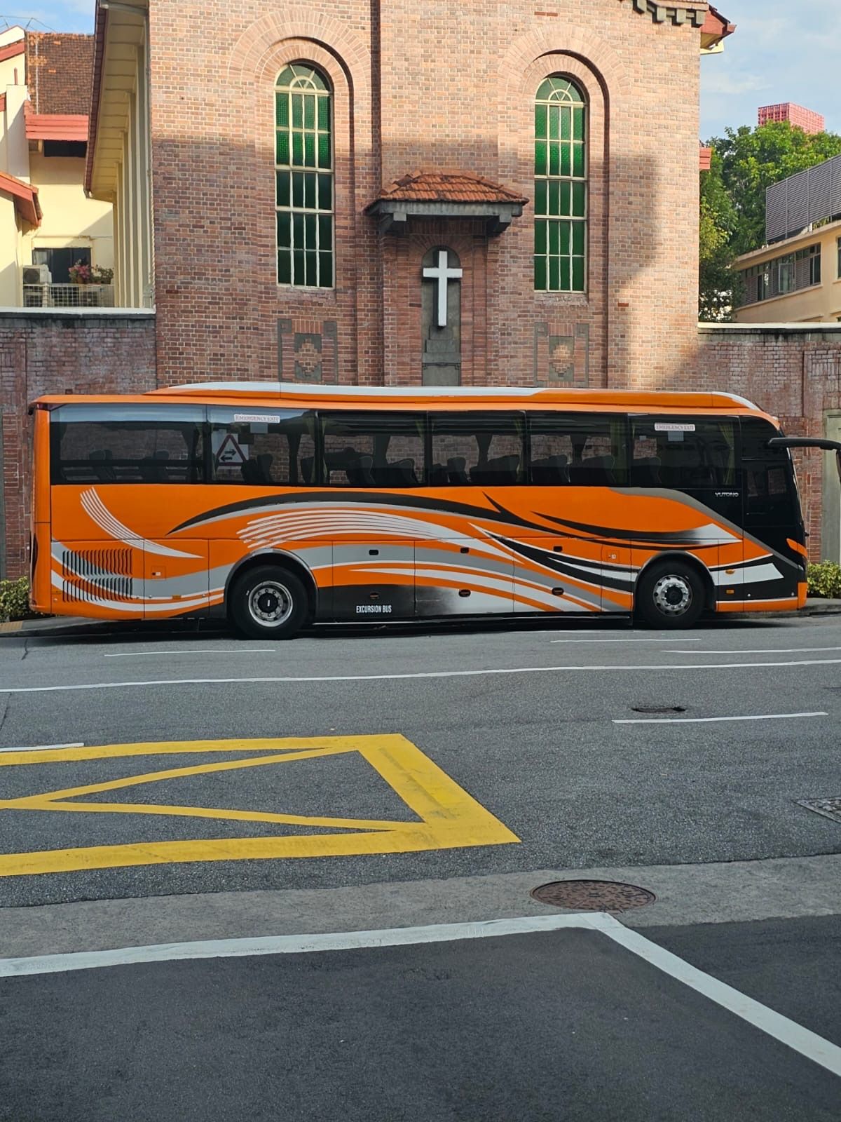 Orange bus with black and silver accents parked on a street in front of a brick building with a cross.