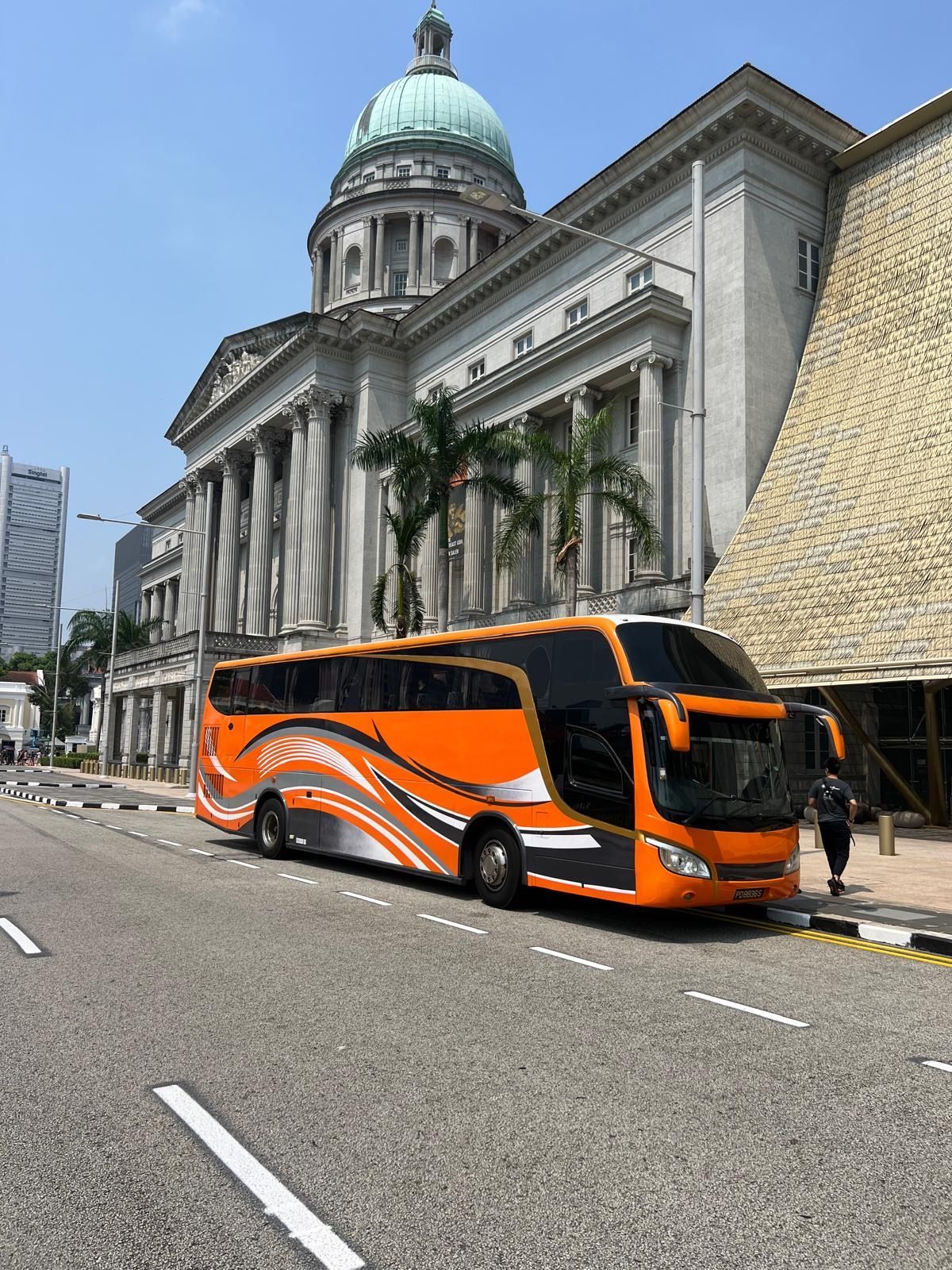Orange tour bus parked in front of a neoclassical building with a dome and tall columns. Palm trees and pedestrians nearby.