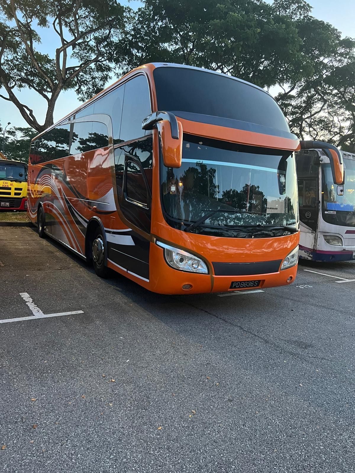 Orange double-decker bus parked in a lot. Other buses and trees are visible in the background.