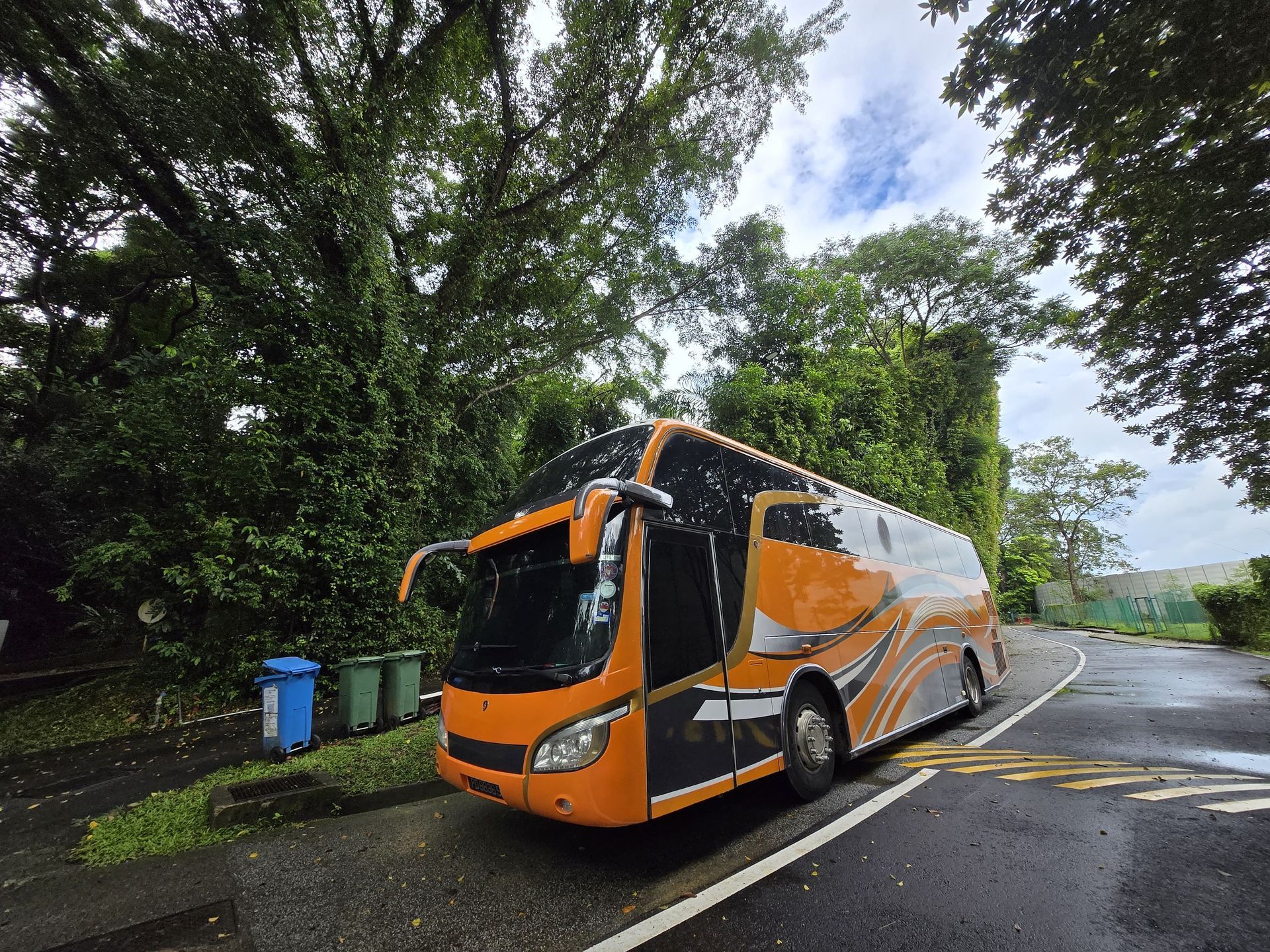 Orange and black bus parked on wet road, surrounded by lush green trees.