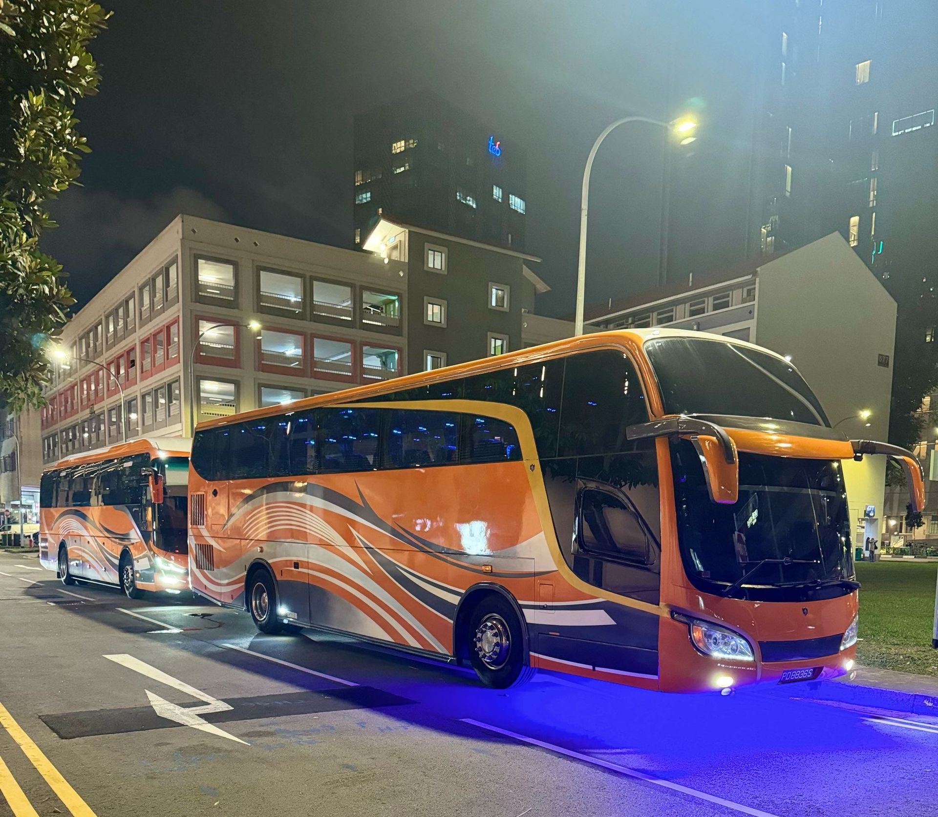 Two orange and white buses parked on a city street at night, with buildings in the background.