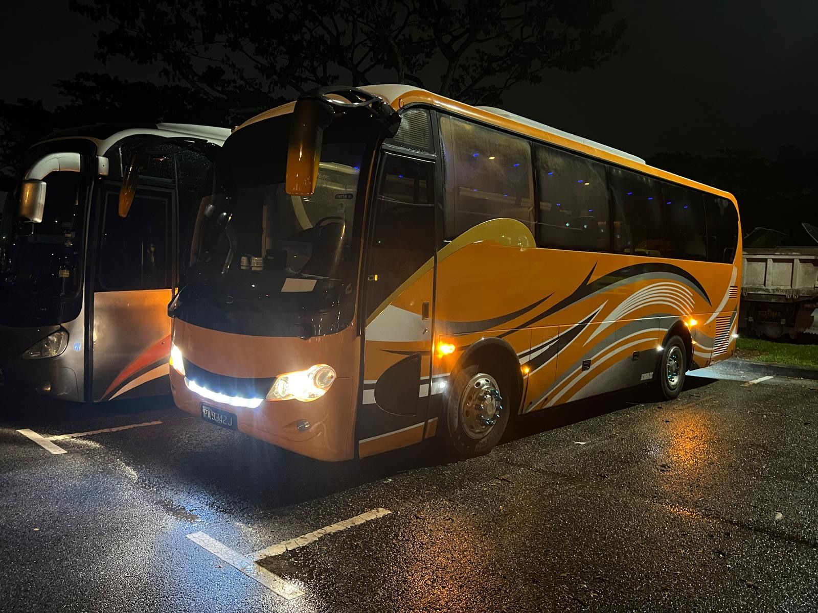 Orange and black bus parked at night, headlights on, next to another bus.