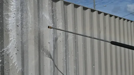 Person using a pressure washer to clean a corrugated metal wall, spraying water.