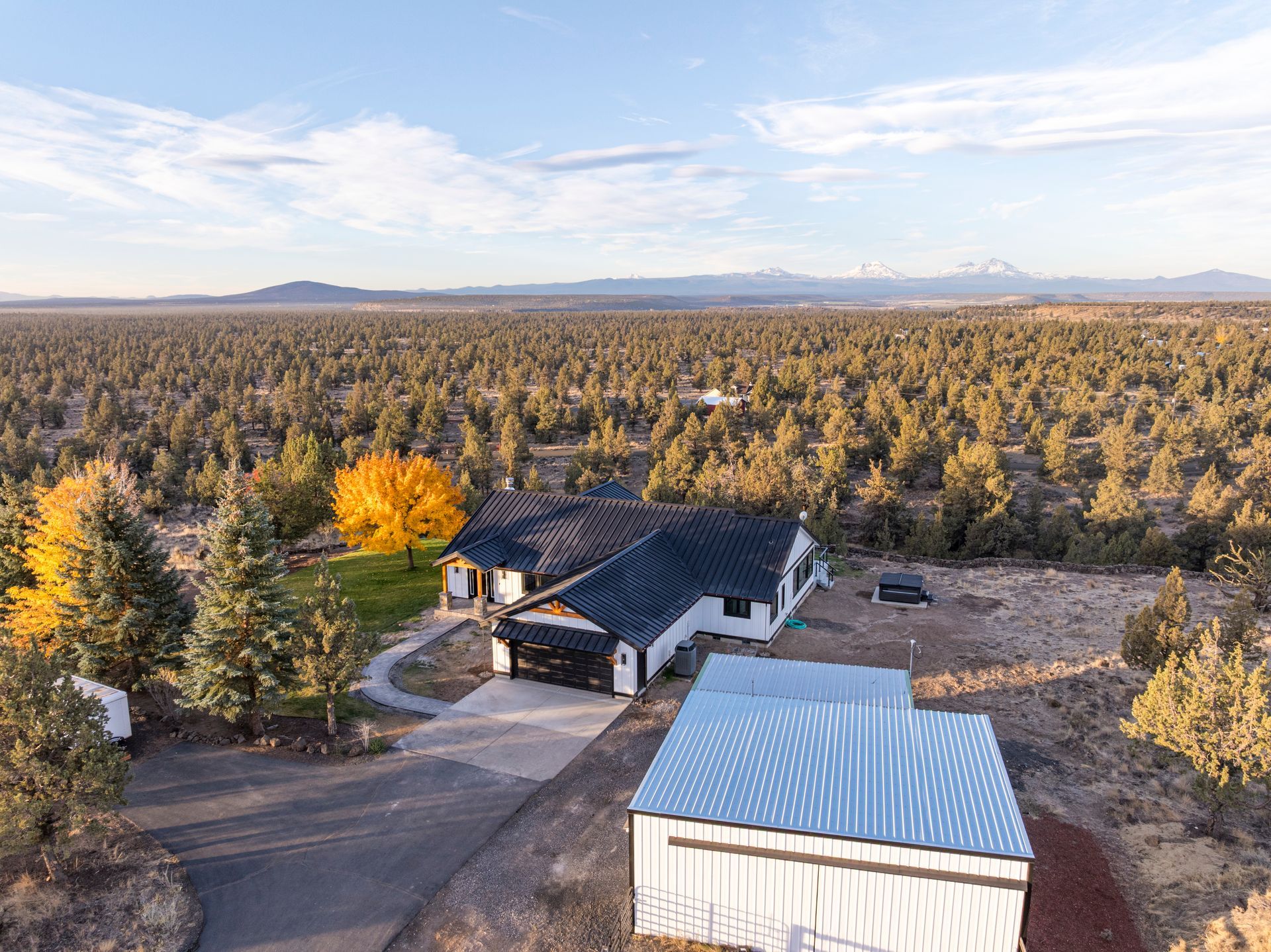 Aerial view of a white house with a black roof and detached garage in a large evergreen forest under a blue sky.