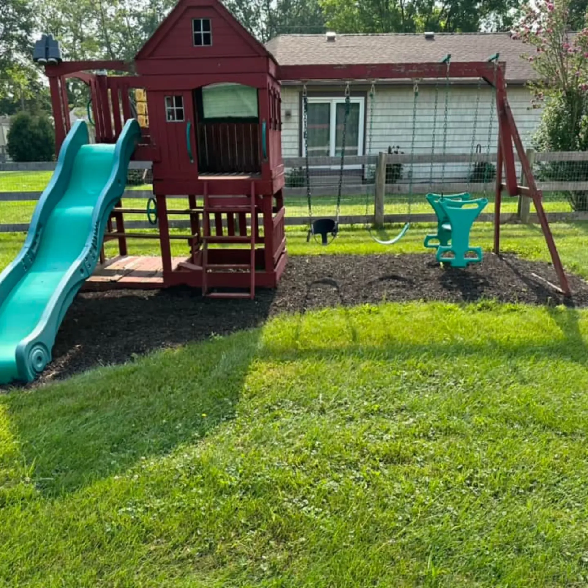 Hall's Hauling -A red playhouse with a green slide and swings in front of a house.