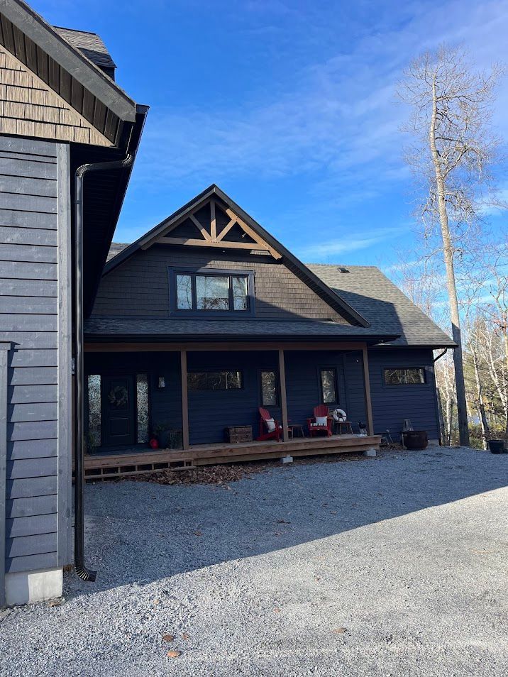Dark blue cabin with porch, brown roof, set against a blue sky, gravel driveway.
