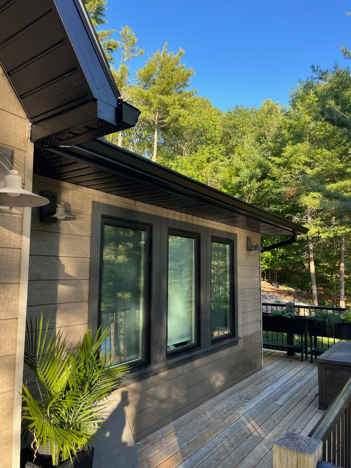 Building exterior with black trim windows, a deck, and lush greenery against a forest backdrop.
