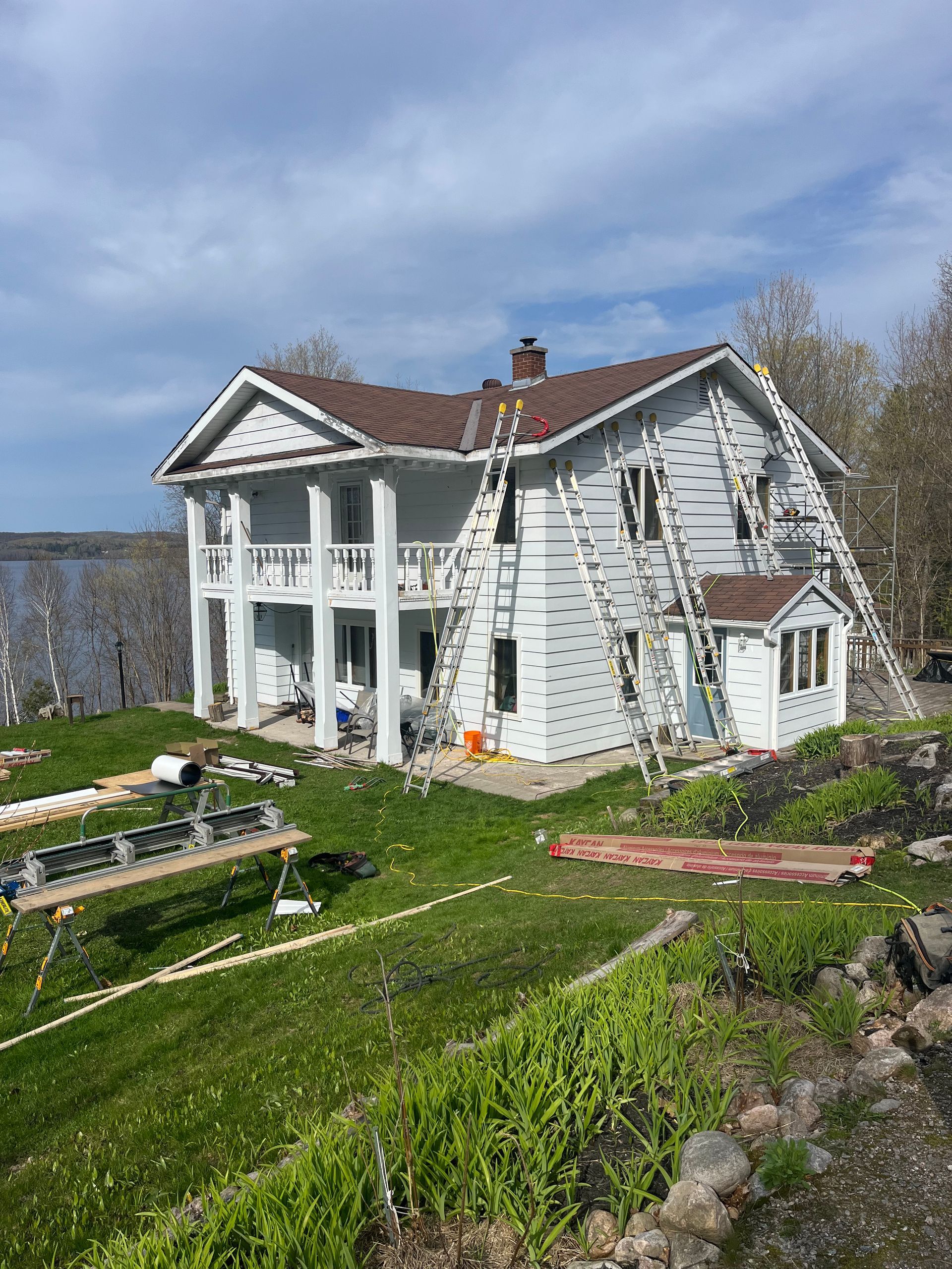 Two-story white house with columns, under renovation, with ladders propped against the siding. Brown roof and green grass in view.