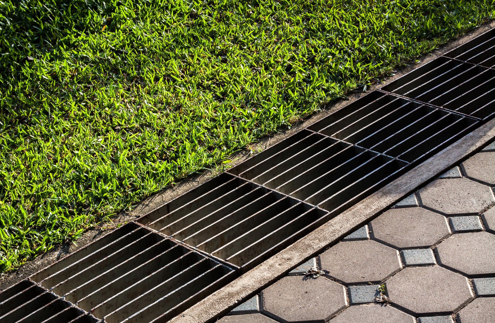 A metal drain on a sidewalk next to a grassy area.