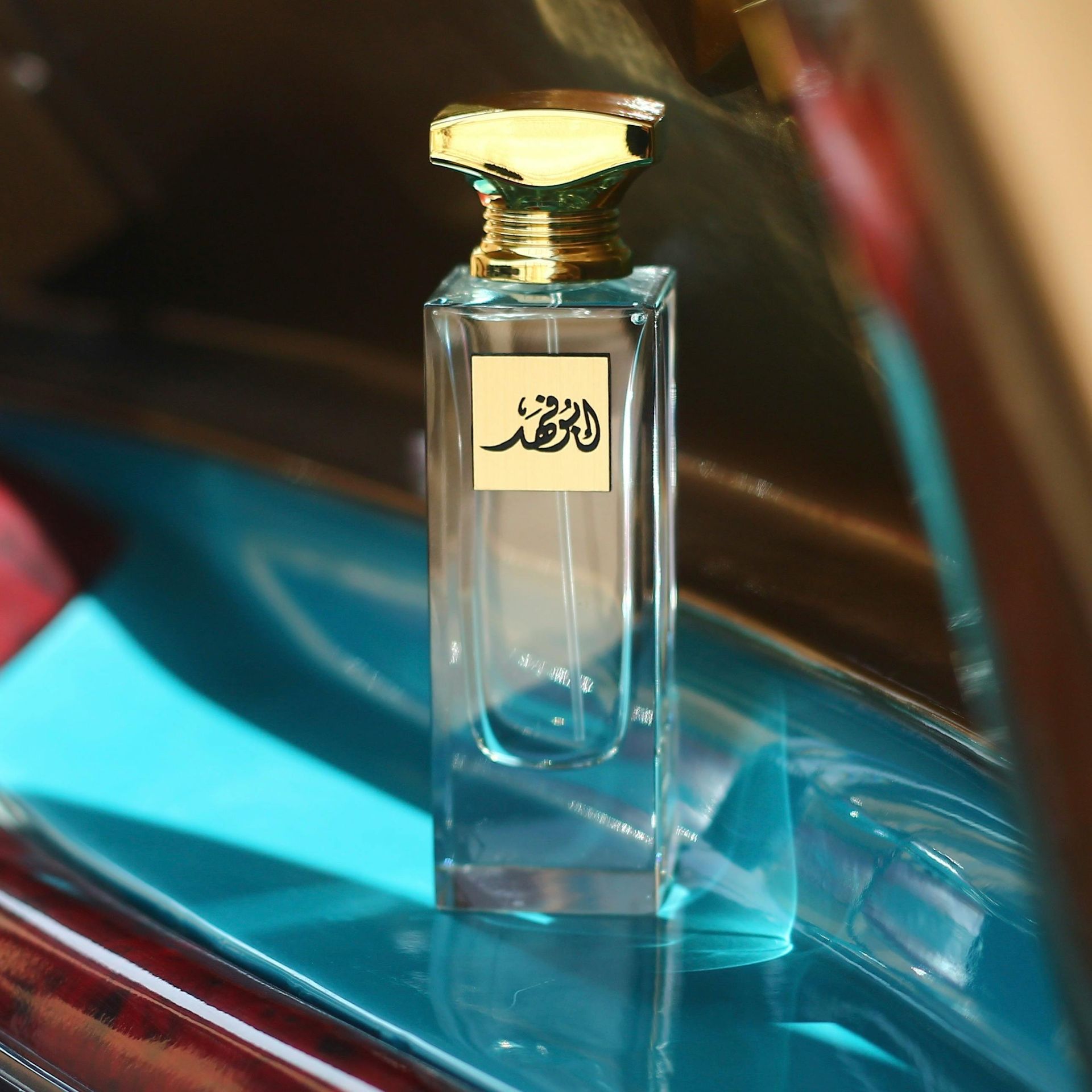 Frasco de perfume, tapón dorado, sobre superficie azul reflectante, con etiqueta en escritura árabe.