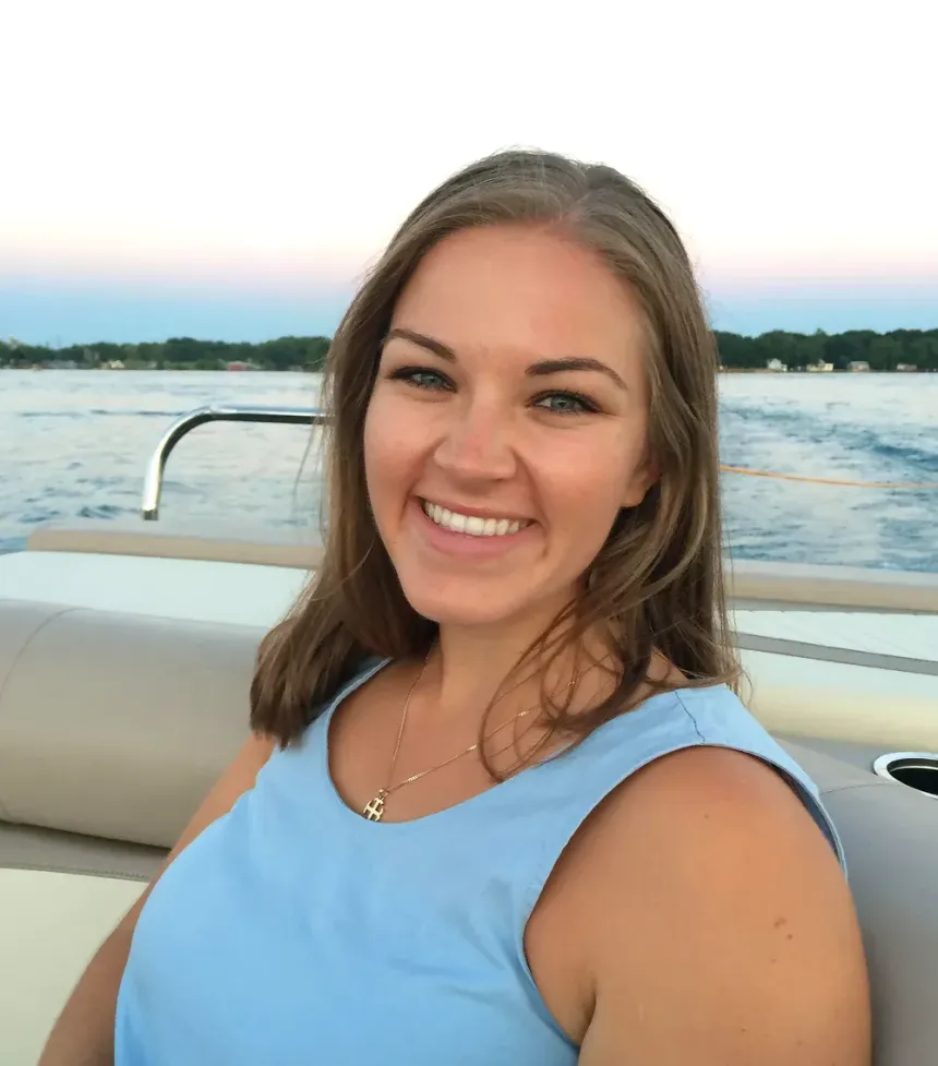Woman with a broad smile, wearing a blue top, on a boat with water and trees in the background.