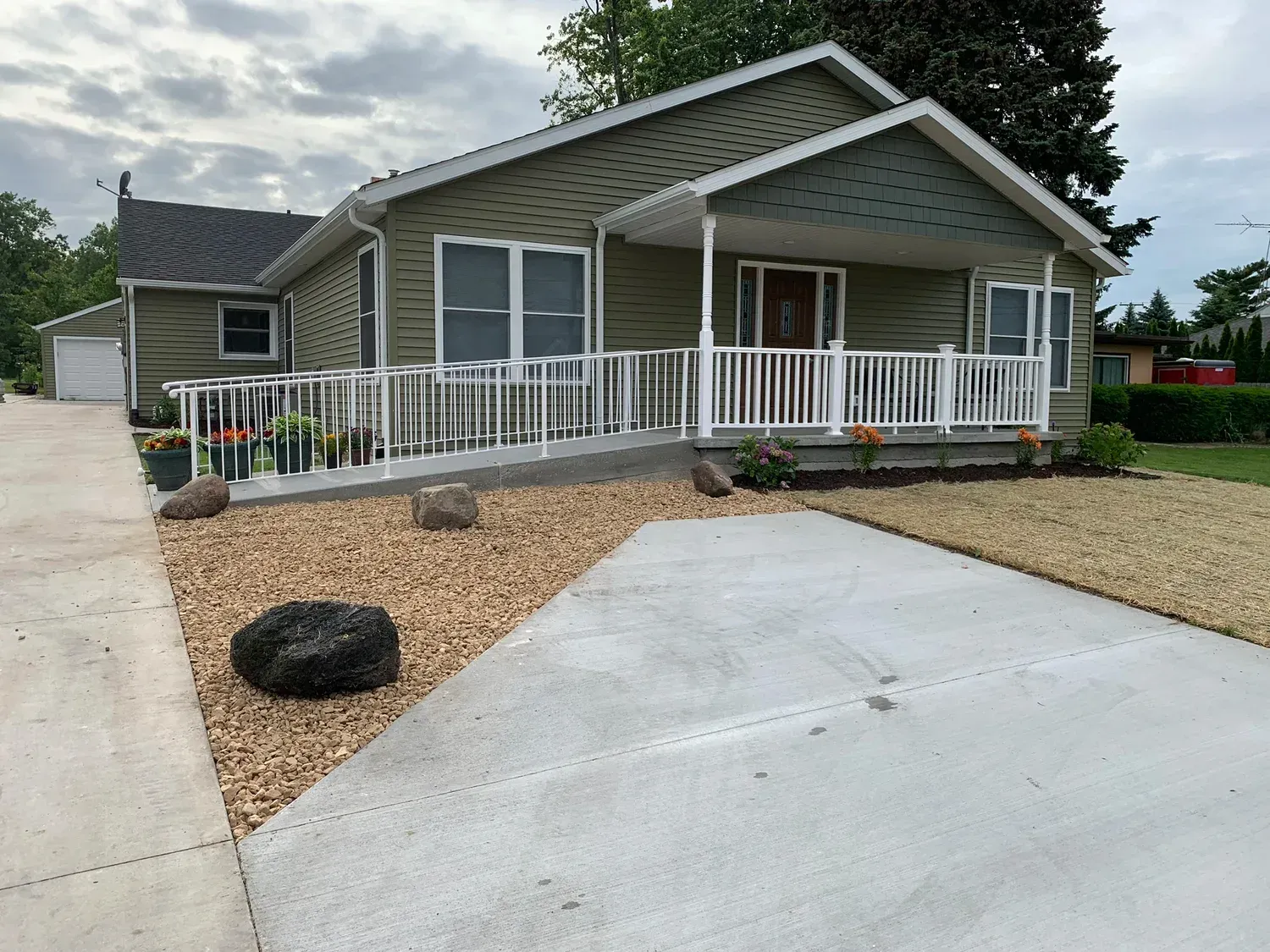 Green house with ramp, white railing, gravel, concrete driveway, and small garden.