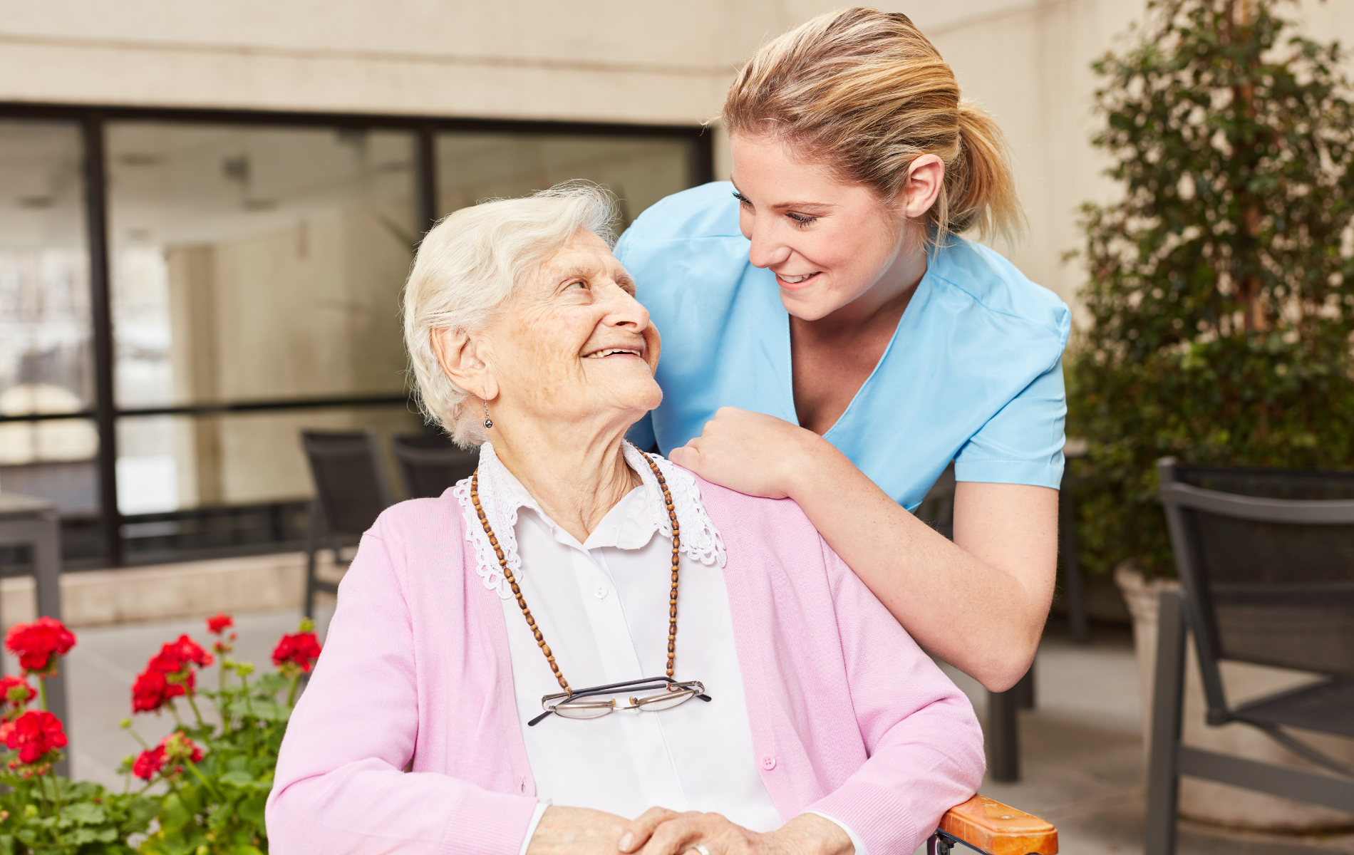 A smiling caregiver in blue scrubs with a hand on the shoulder of a senior in a pink cardigan, outdoors near red flowers.