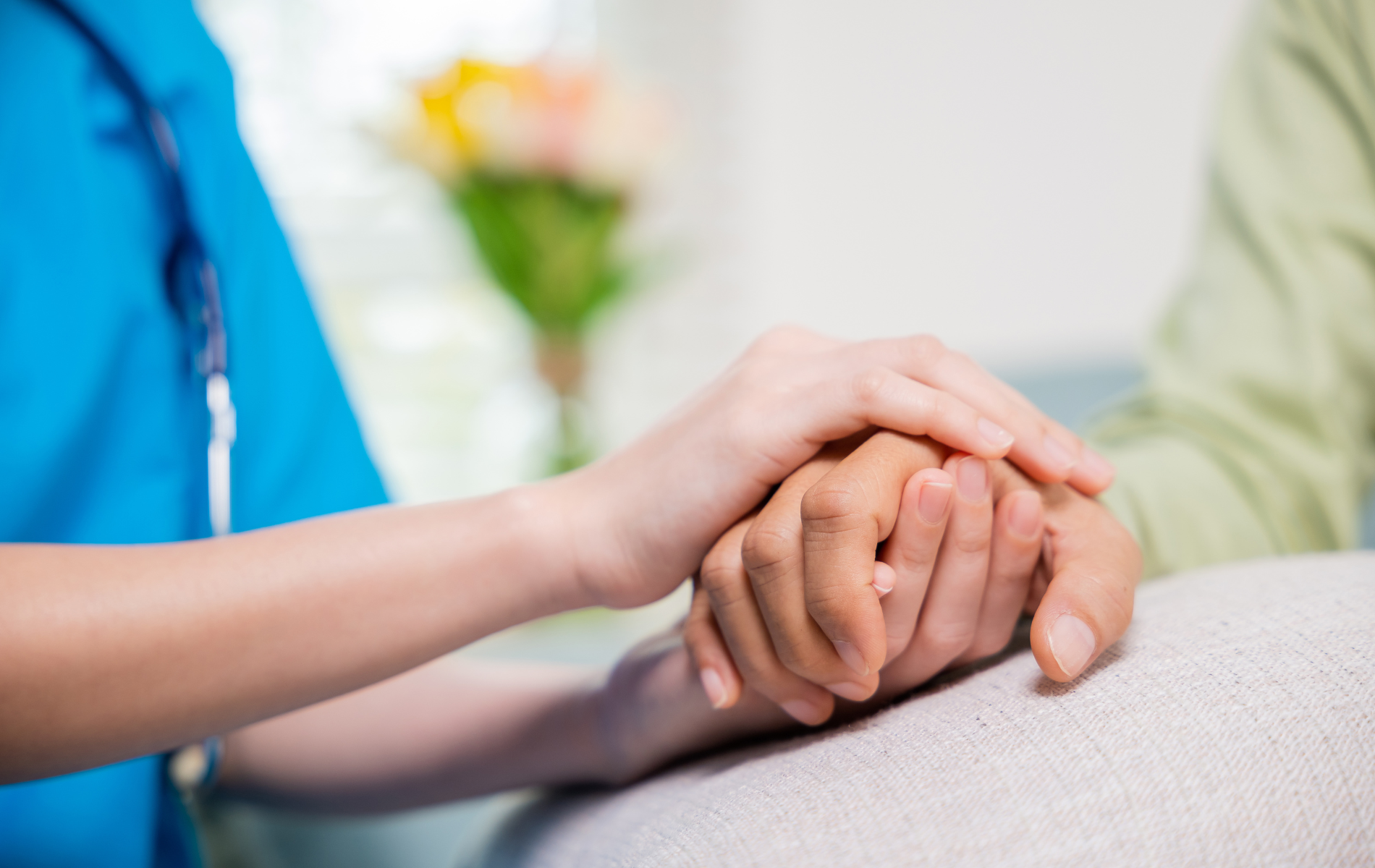 A caregiver in a blue uniform holds a person’s hands gently to provide comfort in a softly lit, warm indoor setting.