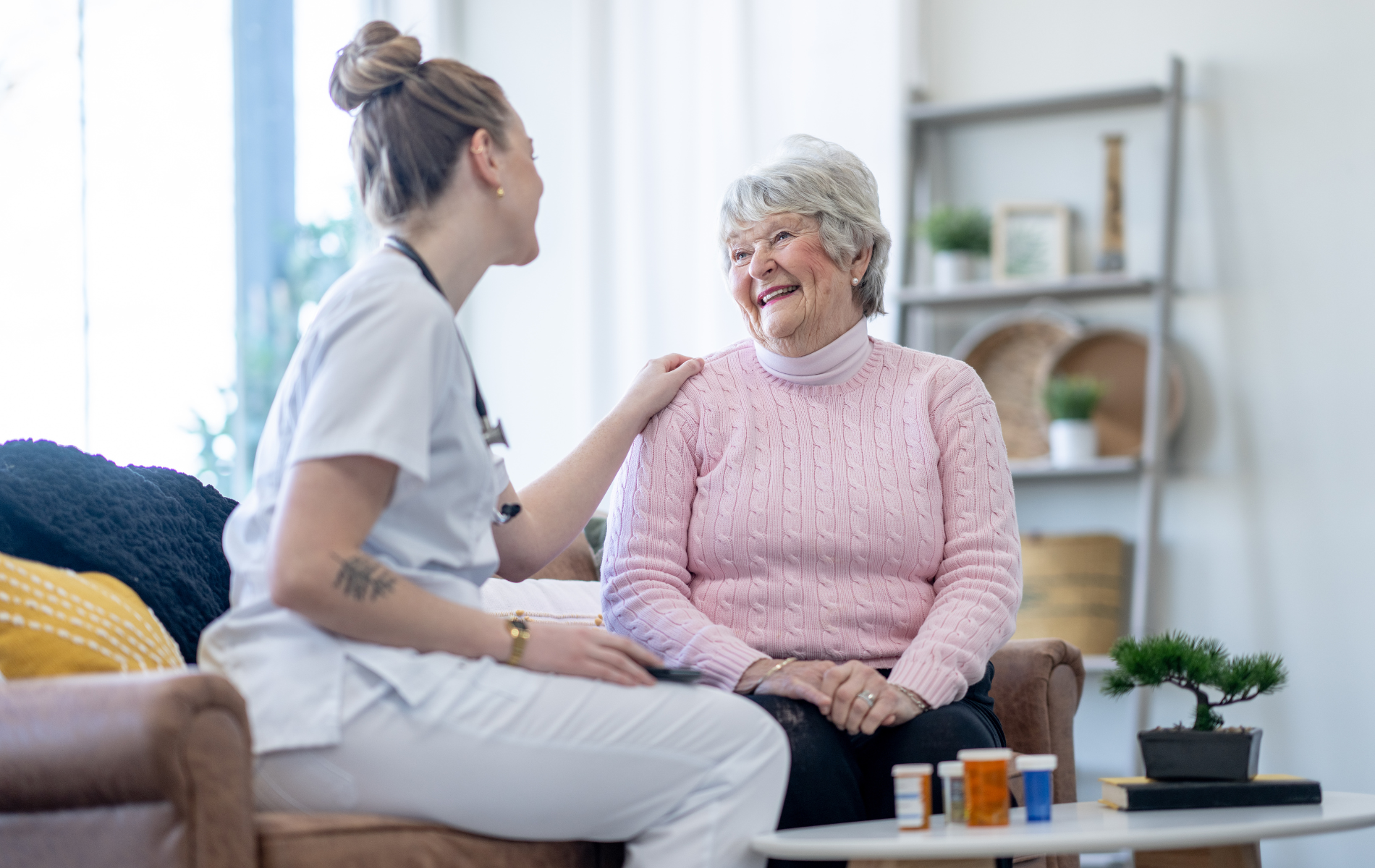 Nurse comforts elderly woman seated on couch, smiling. Bottles of pills in foreground.