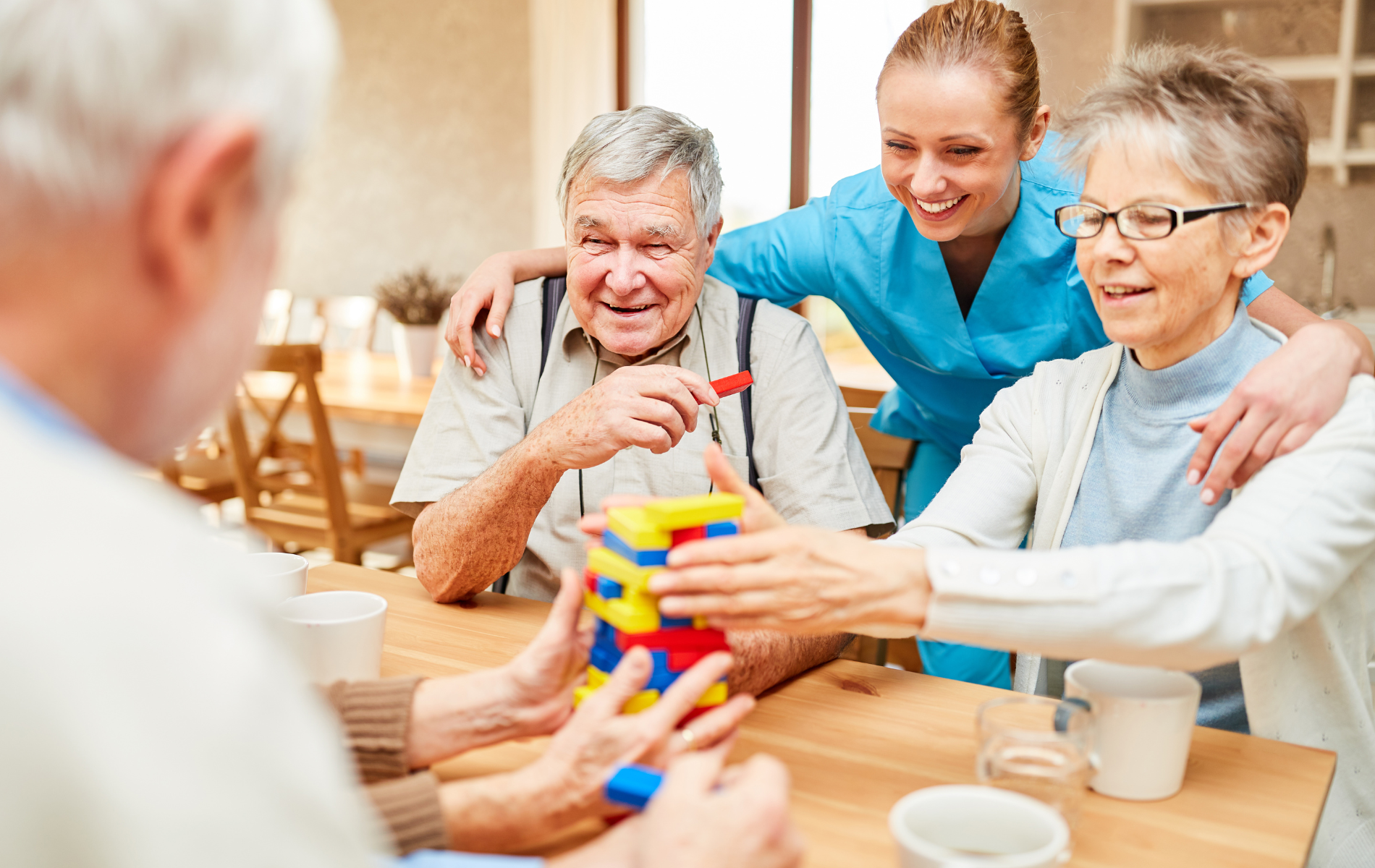 Seniors and caregiver playing with a block puzzle at a table, smiling in a brightly lit room.