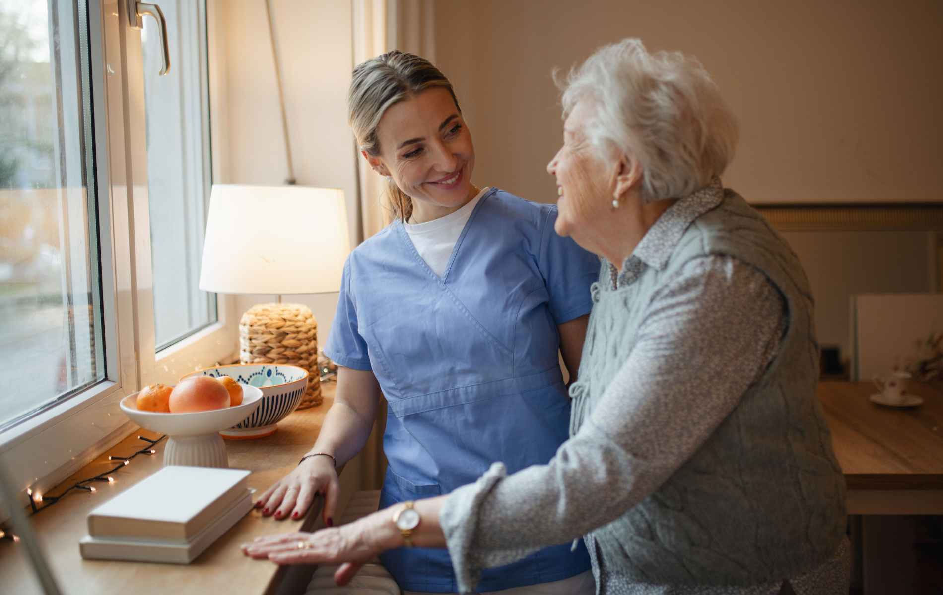 Caregiver in blue scrubs assists an elderly woman. They stand near a window with a friendly, supportive expression.