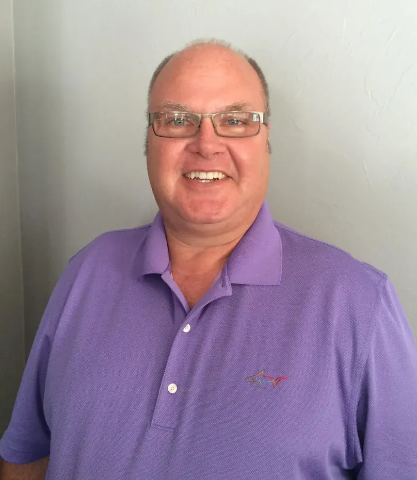 Man with glasses smiles, wearing purple polo shirt, standing in front of a light gray wall.