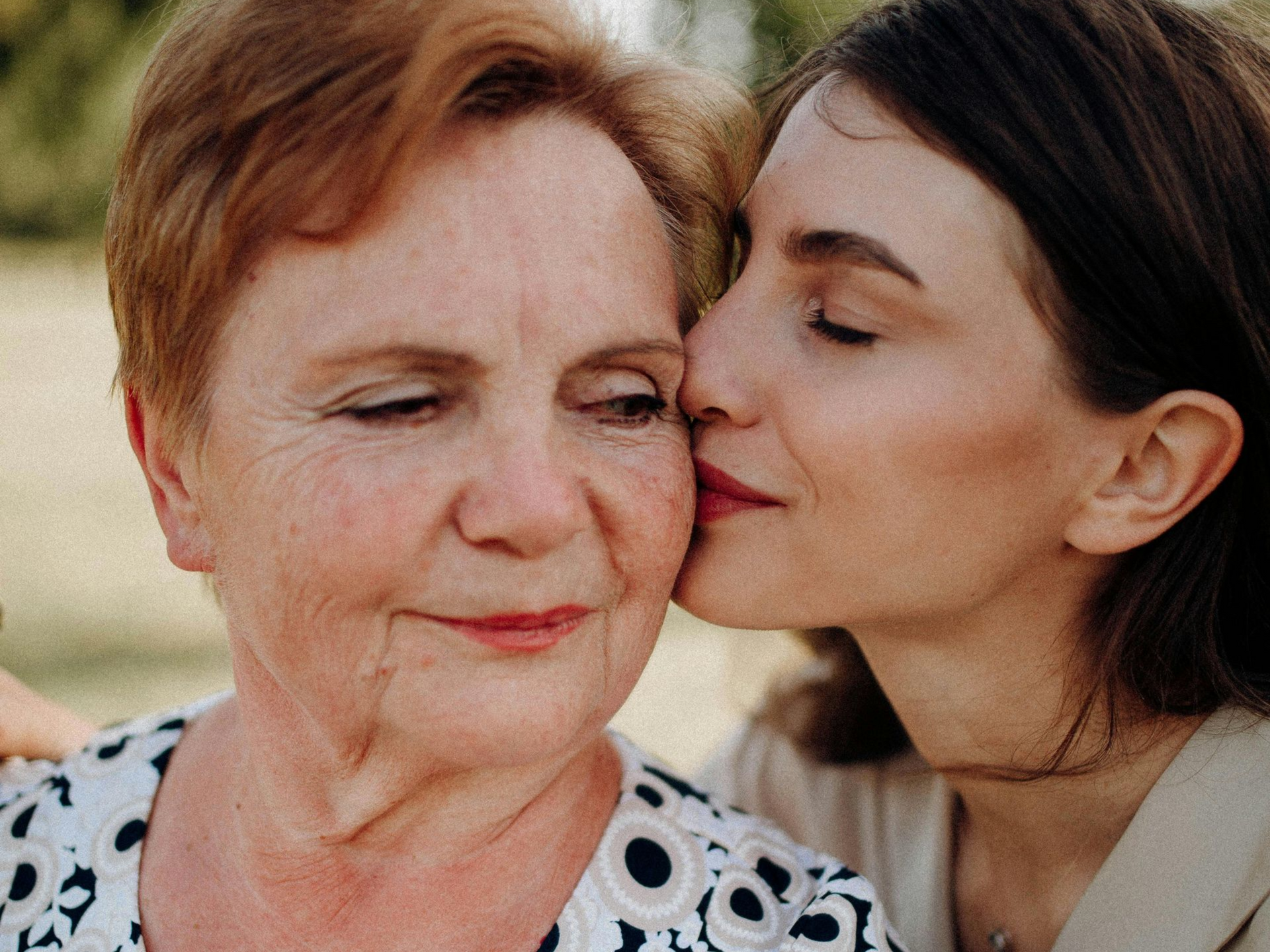 A person gently kissing another person on the cheek outdoors, both with peaceful and warm expressions.