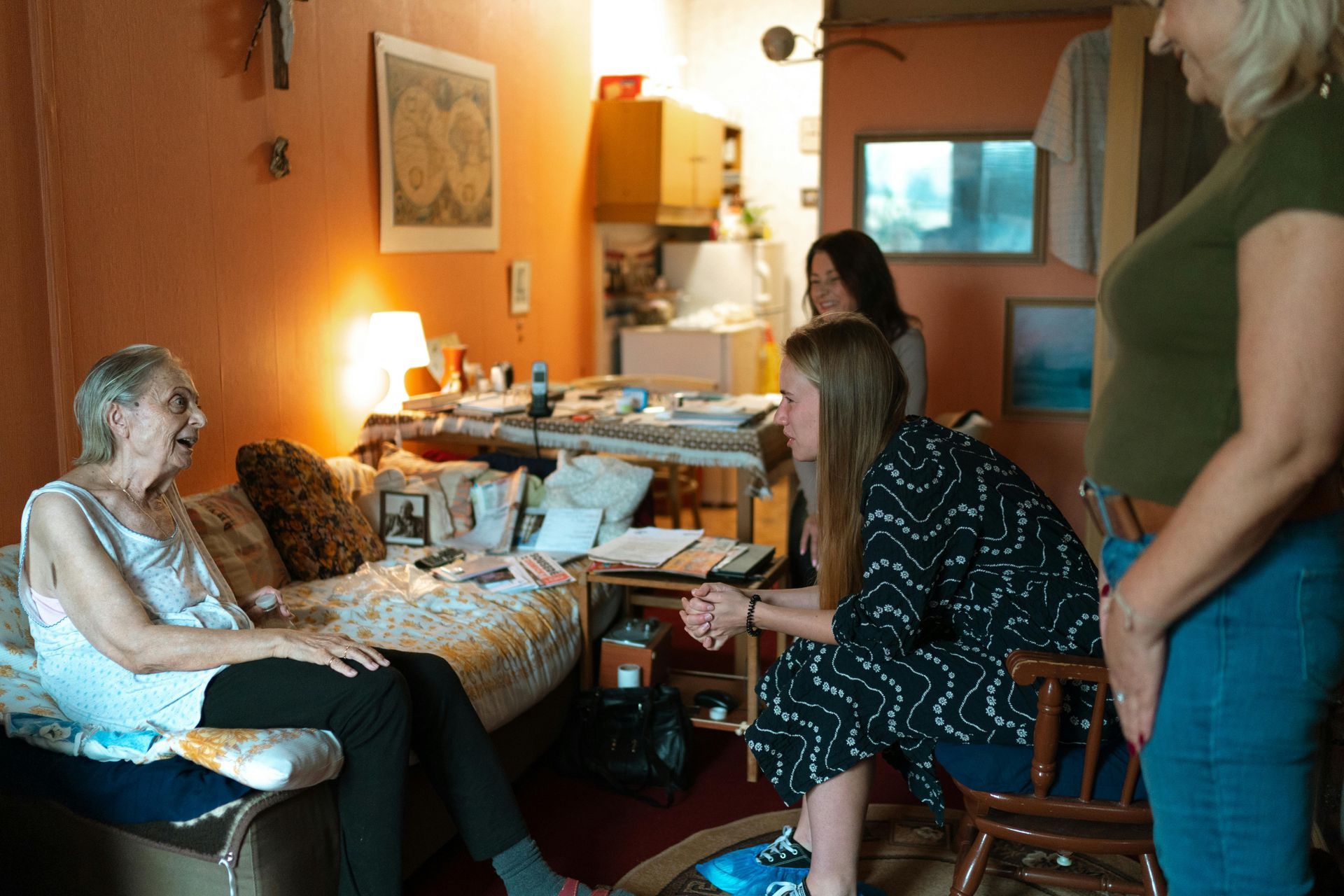 Two caregivers helping a person stand beside a bed in a bright bedroom