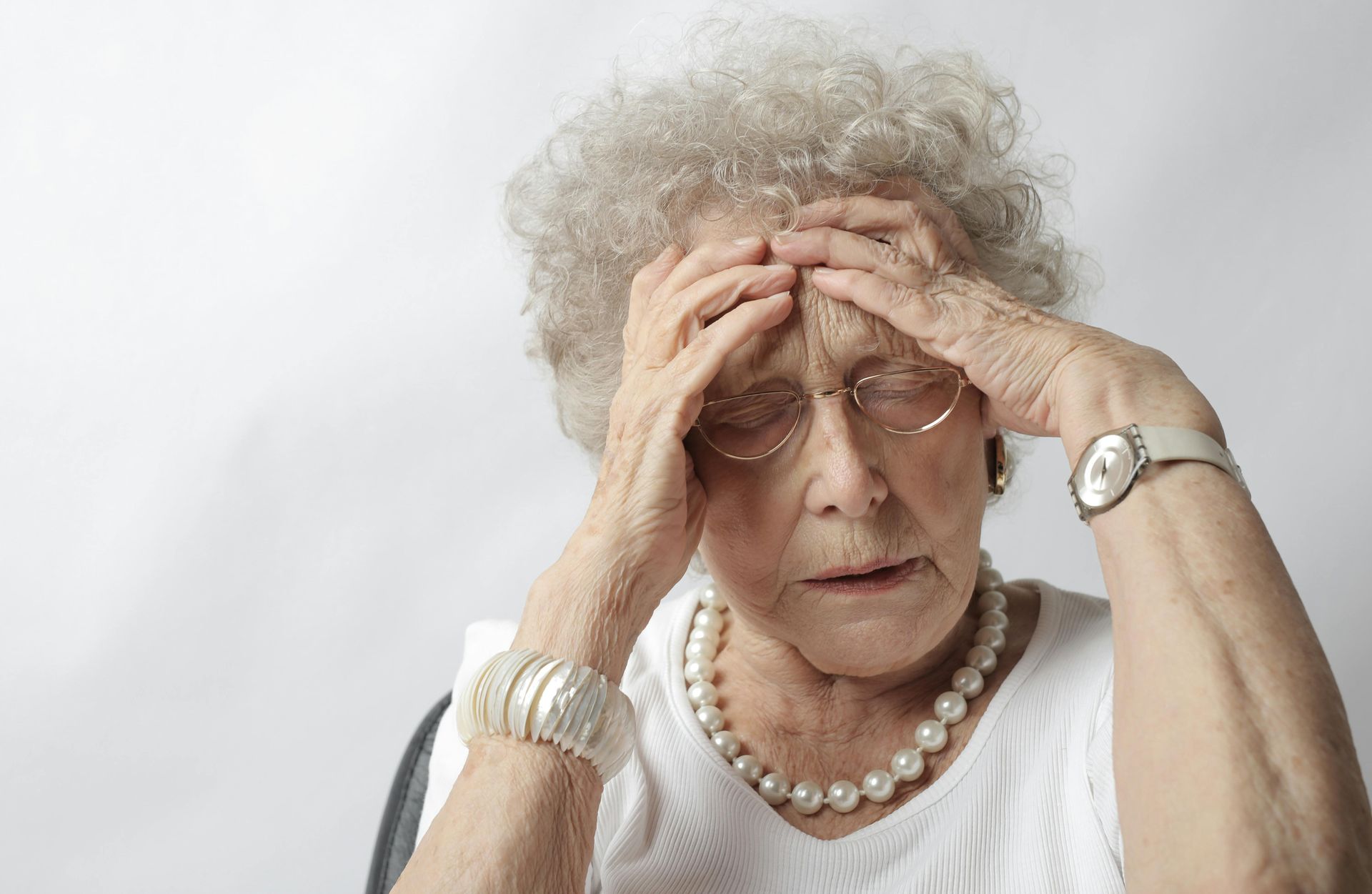 An older person with grey curly hair wearing a white shirt and pearls holding their head with both hands in distress.