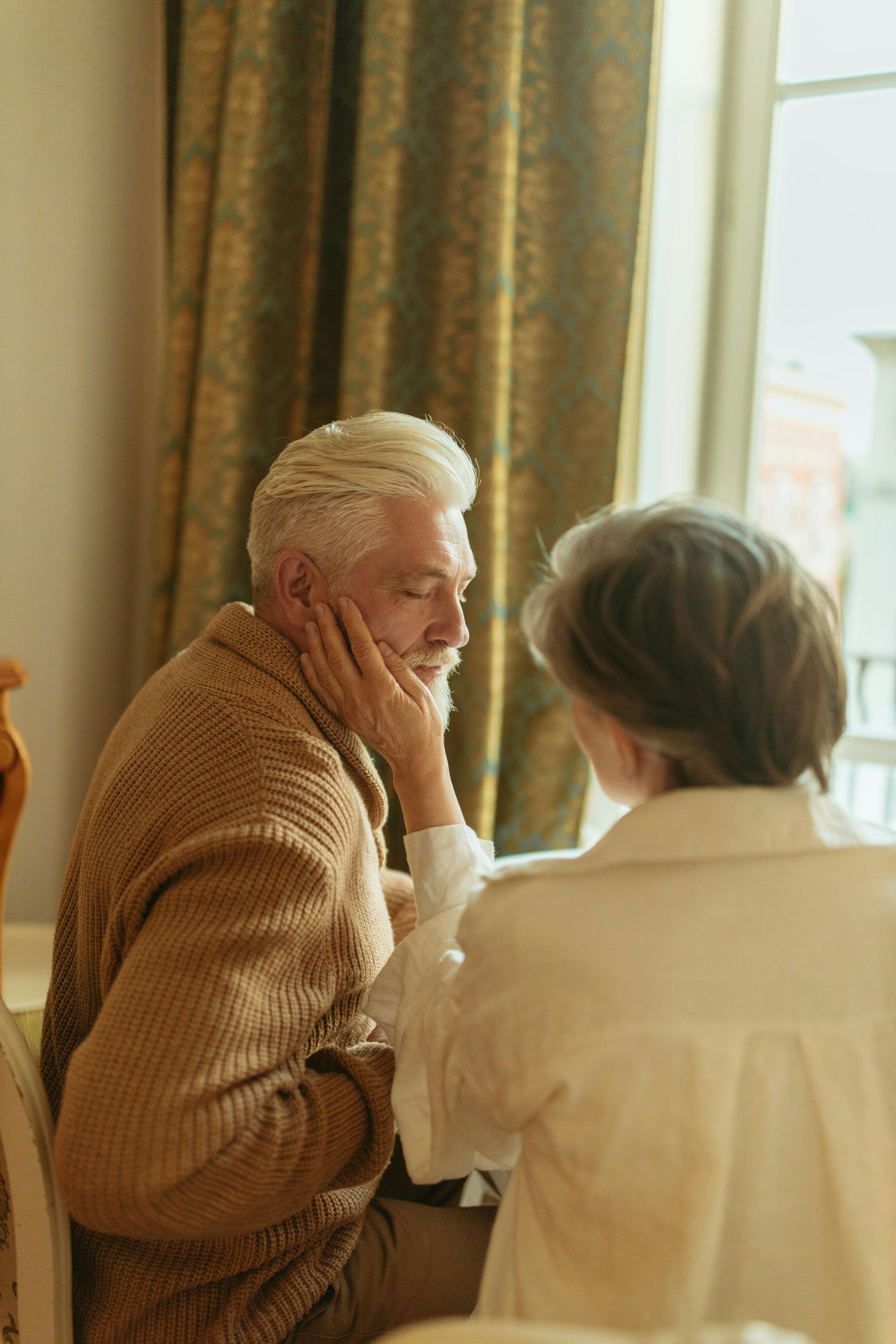 An older person being caressed on the cheek by a person wearing a white shirt; near a window.