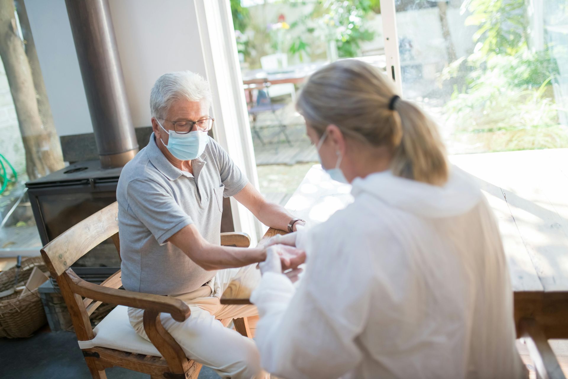 Masked caregiver speaking with a seated patient on a sunny porch