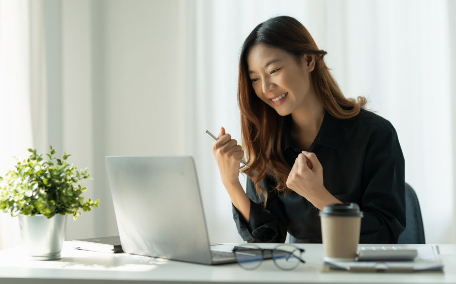 Woman with a joyful expression sitting at a desk with a laptop, holding pen.